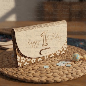 May include: A wooden birthday card with the words "happy 1st birthday" engraved on the front. The card has a floral design and is sitting on a woven mat with confetti and a rattle.