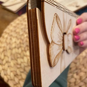 Puede incluir: Una caja de madera de color claro con un dise&ntilde;o de lazo tallado en la parte delantera. La caja est&aacute; sostenida por una persona con esmalte de u&ntilde;as rosa. La caja parece estar hecha de varias capas de madera.