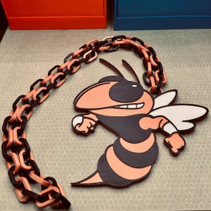 May include: A wooden necklace featuring a stylized cartoon hornet pendant in black, orange, and white. The chain is made of interlocking links in black and orange. The hornet is smiling and has its wings spread.
