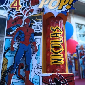 Peut inclure: Une figurine de Spider-Man rouge et bleue se tient devant une case de bande dessinée. La figurine tient un tube rouge et jaune sur lequel est imprimé le mot "NKDLAS". Une bulle de dialogue bleue et jaune avec le mot "POP!" est au-dessus du tube. Une toile d'araignée blanche avec le chiffre "4" en or se trouve dans le coin supérieur gauche de l'image.