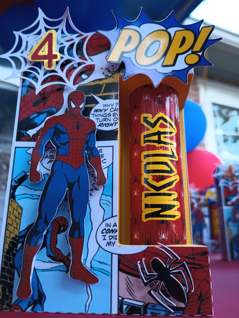 May include: A red and blue Spider-Man figure stands in front of a comic book panel. The figure is holding a red and yellow tube with the word "NKDLAS" printed on it. A blue and yellow speech bubble with the word "POP!" is above the tube. A white spider web with the number "4" in gold is in the top left corner of the image.
