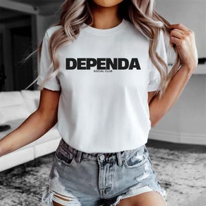 Op de afbeelding: Wit T-shirt met de tekst "DEPENDA SOCIAL CLUB" in vette, zwarte hoofdletters. Het shirt heeft korte mouwen en een ronde hals. Het model draagt een spijkerbroekshort.