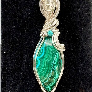 May include: A handcrafted pendant featuring a teardrop-shaped malachite stone with vibrant green and teal patterns. The stone is encased in a silver wire wrap design, with a small turquoise bead accent. The pendant hangs from a black cord.