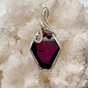 May include: A handcrafted pendant featuring a faceted, hexagonal gemstone with a deep magenta center and black border, encased in a silver wire wrap. Two small, red beads accent the top. The pendant is displayed against a textured, white background.