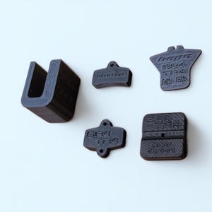 Peut inclure: Cinq objets sombres imprimés en 3D sont disposés sur une surface blanche. Les objets comprennent une pièce en forme de U, une pièce rectangulaire aux bords arrondis et trois pièces avec du texte. Le texte comprend "David Bike Co" et "SR4 TPA".