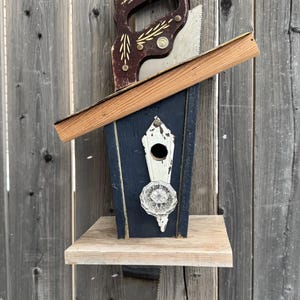 May include: A blue birdhouse with a white door and a glass knob. The birdhouse is attached to a wooden saw with a brown handle.