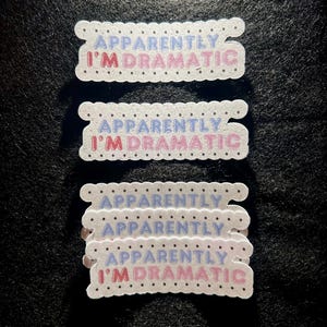 May include: Several white, scalloped-edge stickers with the text "Apparently I'm Dramatic" in blue and pink. The stickers are stacked on a dark surface.