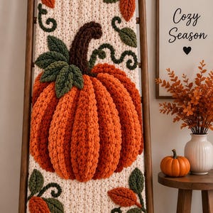 Könnte beinhalten: Eine cremefarbene, grob gestrickte Decke mit einem großen, strukturierten orangefarbenen Kürbis, einem dunkelbraunen Stiel und grünen Blättern. Das Design enthält kleinere orange und grüne Blattakzente, die auf der weichen Wolle verteilt sind.