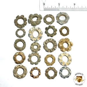 May include: A collection of antique, weathered metal rings and decorative pieces, some with a floral or cog-like design. The items vary in size and color, ranging from gray to brown. A ruler is in the background.