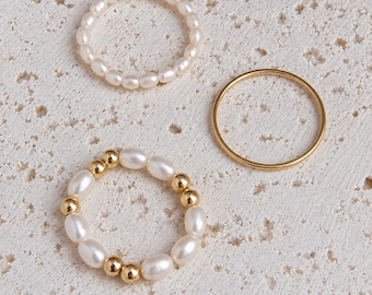 May include: A set of three rings including one band made of small white pearls with a single gold bead, one plain thin gold band, and one band featuring alternating white oval pearls and small gold beads arranged in a circular pattern.