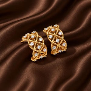 May include: A pair of gold hoop earrings featuring a braided rope design with embedded clear round stones. The earrings have a curved, open-work structure that showcases the intricate metalwork and the sparkling stones set throughout.