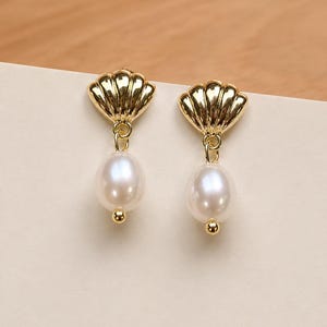 May include: A pair of gold-toned earrings featuring a polished shell-shaped stud at the top. A single white oval-shaped pearl hangs from each shell, finished with a small gold-toned bead at the bottom of each pearl, creating a dangling design.