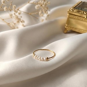 May include: A thin gold ring featuring a row of five small, round white pearls spaced by tiny gold beads. The band is constructed from a slender gold wire, creating a delicate circular shape that rests on a soft, draped off-white fabric.