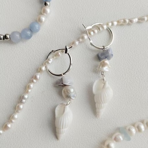 May include: A pair of silver hoop earrings featuring white spiral seashell charms, small grey stone beads, and white pearls. The earrings are displayed alongside strands of white pearls interspersed with light blue beads.