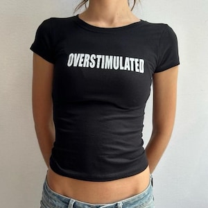 Puede incluir: Camiseta negra de manga corta con la palabra "OVERSTIMULATED" en blanco en el pecho. La camiseta ajustada se combina con vaqueros azules. El look general es casual y moderno.