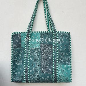 Puede incluir: Un bolso tote acolchado con un diseño de patchwork de patrones de bandana de cachemira en verde oscuro y verde azulado. El bolso tiene dos asas a juego y bordes a rayas verdes y blancas. El texto @BohoCraftJaipur está impreso en blanco.