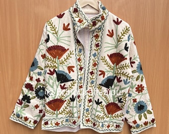 Può includere: Una giacca color crema con maniche lunghe, collo alto e chiusura frontale con zip. Il capo è decorato con intricati ricami floreali nelle tonalità del rosso, blu, verde e nero. La fodera interna presenta delle sottili righe rosse.