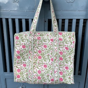 Puede incluir: Un bolso tote acolchado con un estampado floral de rosas rosas y hojas verdes sobre un fondo blanco. El bolso tiene asas largas y un ribete a rayas verdes y blancas. El texto "jaipurcraftstextiles" está impreso en el bolso.