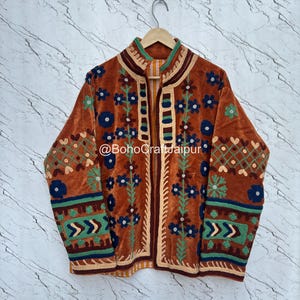 May include: A rust-colored velvet jacket featuring intricate embroidery in shades of blue, green, and cream. The garment displays floral patterns, geometric borders, and a high collar with a front opening. The text @BohoCraftJaipur is visible.