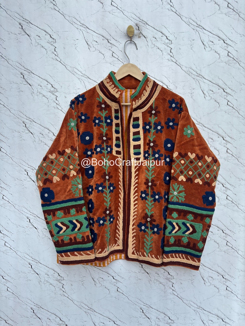 May include: A rust-colored velvet jacket featuring intricate embroidery in shades of blue, green, and cream. The garment displays floral patterns, geometric borders, and a high collar with a front opening. The text @BohoCraftJaipur is visible.