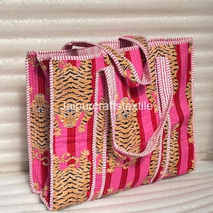 Puede incluir: Bolso tote rosa y rojo con estampado de rayas de tigre. El bolso presenta un diseño acolchado con rayas verticales e ilustraciones de tigres. Las asas son rosas y blancas. El texto "jaipurcraftstextile" es visible.