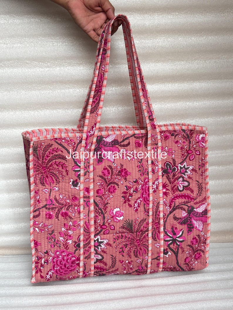 May include: A pink tote bag with a floral pattern in shades of pink and magenta. The bag has long handles and a rectangular shape. The text "jaipurcraftstextile" is visible on the bag.