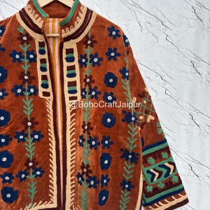 May include: A rust-colored jacket featuring intricate dark blue and green floral embroidery, a high collar, and a striped inner lining. The garment displays the text BohoCraftJaipur and includes geometric patterns on the sleeves and front edges.