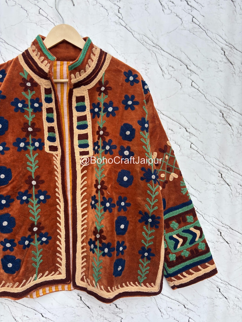 May include: A rust-colored jacket featuring intricate dark blue and green floral embroidery, a high collar, and a striped inner lining. The garment displays the text BohoCraftJaipur and includes geometric patterns on the sleeves and front edges.