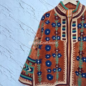 May include: A rust-colored velvet jacket featuring intricate floral embroidery in blue, green, and cream. The garment displays a high collar, long sleeves with geometric patterns, and the text @BohoCraftJaipur printed across the center front.