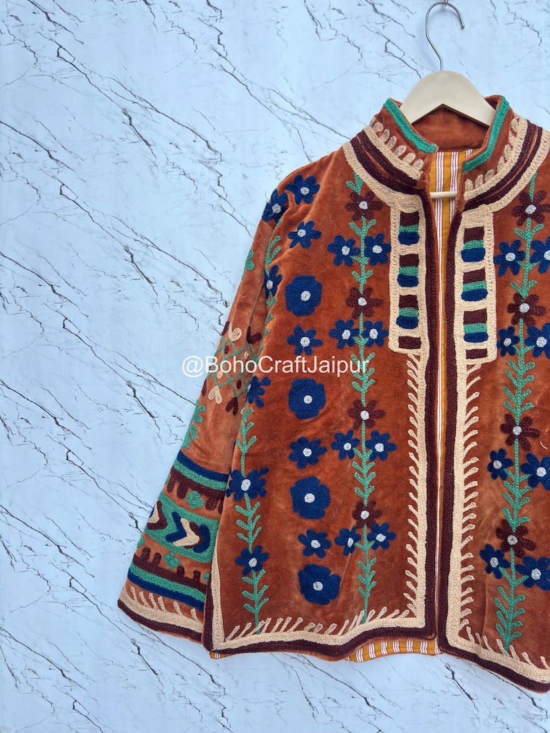 May include: A rust-colored velvet jacket featuring intricate floral embroidery in blue, green, and cream. The garment displays a high collar, long sleeves with geometric patterns, and the text @BohoCraftJaipur printed across the center front.