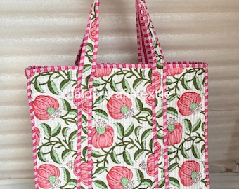 May include: A quilted tote bag with a floral design. The bag has a white base with pink flowers and green leaves. The handles and trim are pink and white striped. The bag is held by a hand.