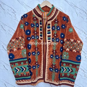 May include: A burnt orange velvet jacket featuring intricate multi-colored floral and geometric embroidery in shades of blue, green, and cream. The garment has a high collar, long sleeves, and the text @BohoCraftJaipur printed on the front.