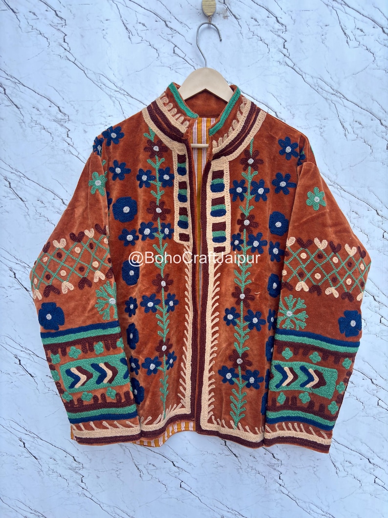 May include: A burnt orange velvet jacket featuring intricate multi-colored floral and geometric embroidery in shades of blue, green, and cream. The garment has a high collar, long sleeves, and the text @BohoCraftJaipur printed on the front.