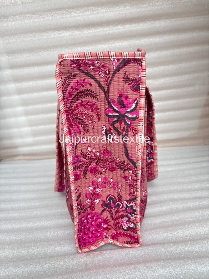 May include: A quilted tote bag with a floral pattern in shades of pink and magenta. The bag features a repeating design of stylized flowers and foliage. The edges are trimmed with a striped fabric in pink and white. The text "jaipurcrafttextile" is visible.