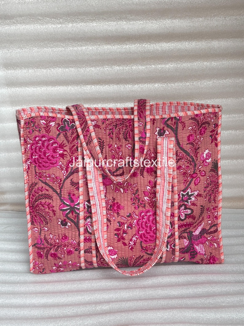 May include: A quilted tote bag with a floral pattern in shades of pink and magenta. The bag has two handles and a rectangular shape. The text "Jamurcraftstextile" is visible on the bag.