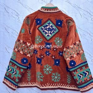 May include: A rust-colored jacket featuring intricate multi-colored embroidery with floral and geometric patterns, a high collar, and long sleeves. The text @BohoCraftJaipur is printed across the center back of the garment.