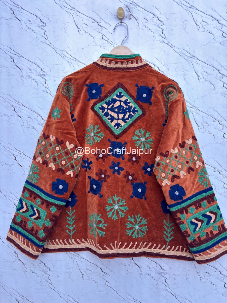 May include: A rust-colored jacket featuring intricate multi-colored embroidery with floral and geometric patterns, a high collar, and long sleeves. The text @BohoCraftJaipur is printed across the center back of the garment.