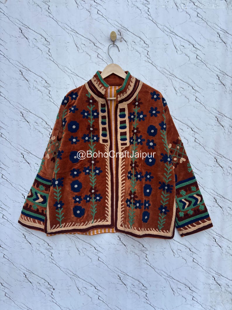 May include: A rust-colored jacket featuring intricate embroidery in dark blue, green, and cream. The design includes floral patterns, geometric borders, and a high collar. The text @BohoCraftJaipur is printed across the center of the garment.