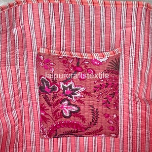 May include: Close-up of a pink and white striped fabric with a floral pocket. The pocket features a dark pink floral pattern with the text "jaipurcraftstextile". The fabric has a quilted texture.