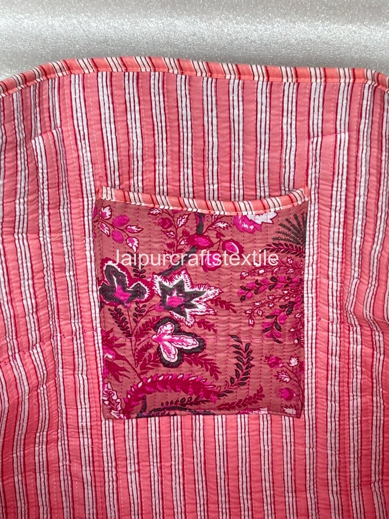 May include: Close-up of a pink and white striped fabric with a floral pocket. The pocket features a dark pink floral pattern with the text "jaipurcraftstextile". The fabric has a quilted texture.