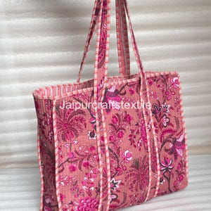 May include: A quilted tote bag with a floral pattern in shades of pink and magenta. The bag has long handles and a rectangular shape. The text "Jaipurcrafttextile" is visible on the bag.