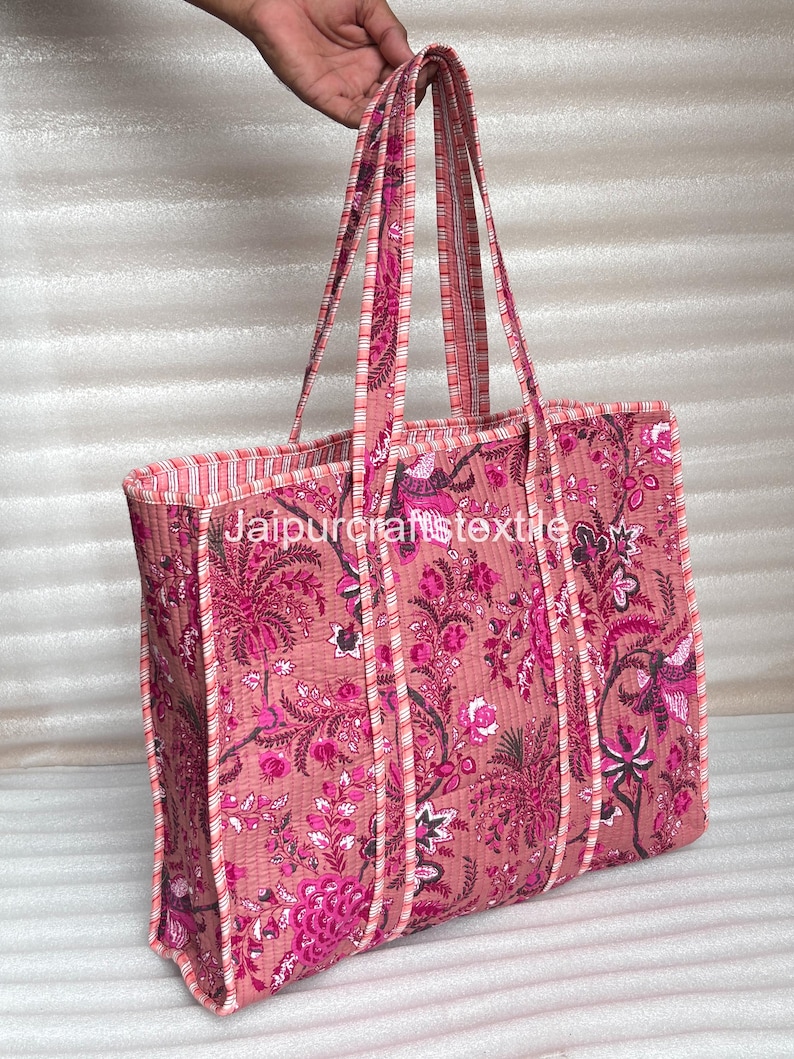 May include: A quilted tote bag with a floral pattern in shades of pink and magenta. The bag has long handles and a rectangular shape. The text "Jaipurcrafttextile" is visible on the bag.