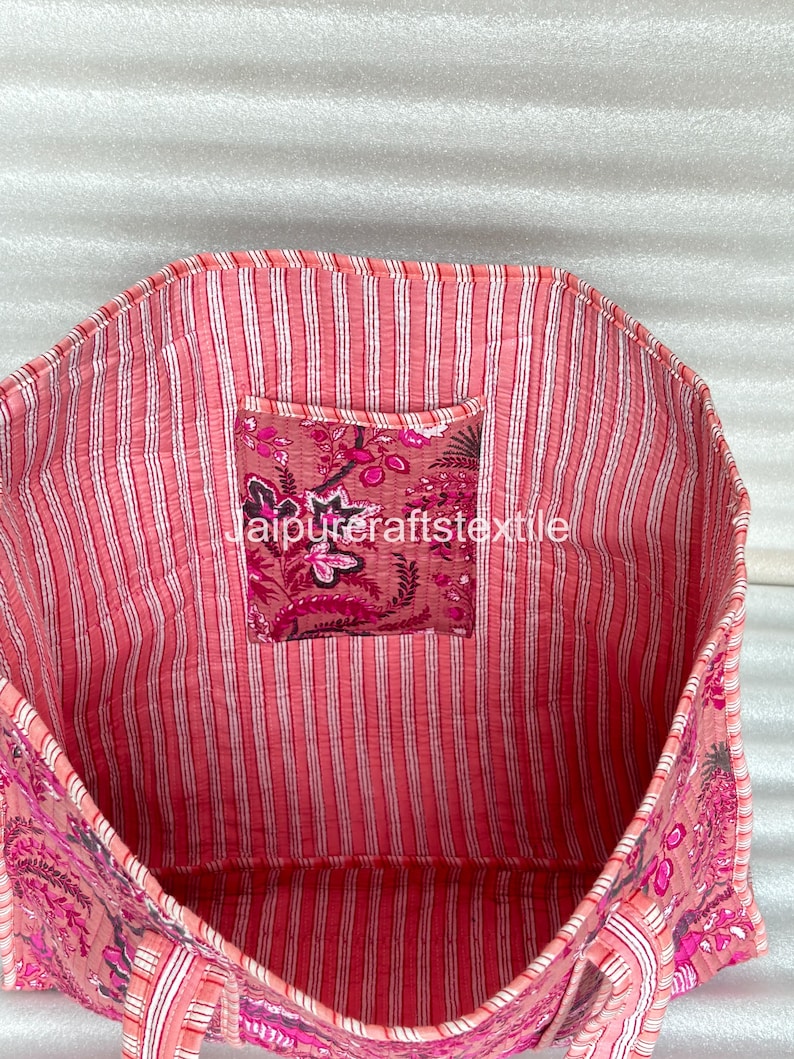 May include: Open view of a pink tote bag with a floral pattern and striped lining. The bag has a small pocket with a floral design and the text "jaipurcraftstextile". The bag is made of a soft, quilted fabric.