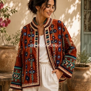 May include: A rust-colored velvet jacket featuring a mandarin collar and intricate floral embroidery in blue, green, and cream. The garment displays geometric patterns on the sleeves and front panels, with the text @BohoCraftJaipur visible.