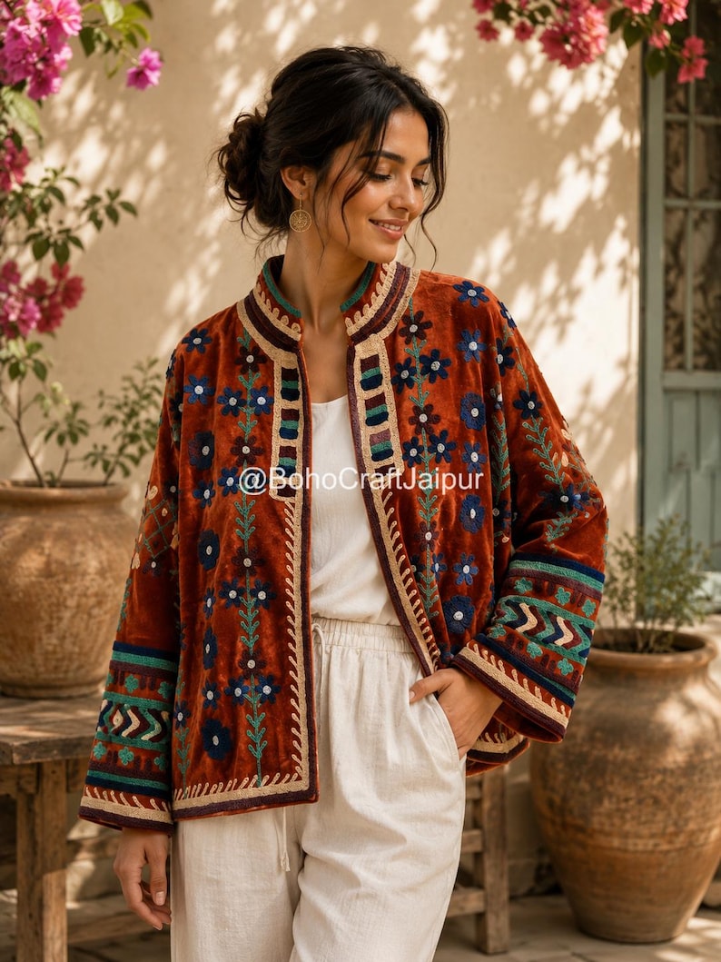 May include: A rust-colored velvet jacket featuring a mandarin collar and intricate floral embroidery in blue, green, and cream. The garment displays geometric patterns on the sleeves and front panels, with the text @BohoCraftJaipur visible.