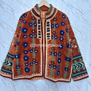 May include: A burnt orange velvet jacket featuring intricate floral embroidery in shades of blue, green, and cream. The garment has a high collar, long sleeves with geometric patterns, and the text @BohoCraftJaipur printed across the center front.