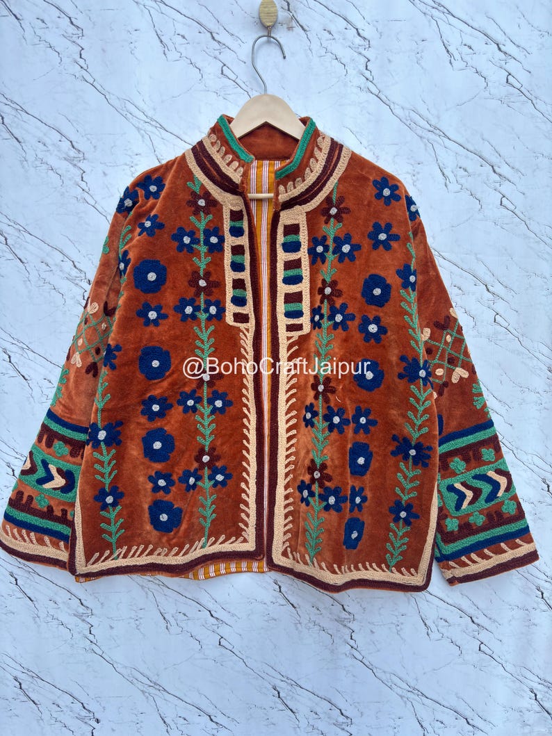 May include: A burnt orange velvet jacket featuring intricate floral embroidery in shades of blue, green, and cream. The garment has a high collar, long sleeves with geometric patterns, and the text @BohoCraftJaipur printed across the center front.