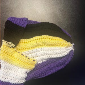 Könnte beinhalten: Eine gehäkelte Mütze in den Farben der nicht-binären Pride-Flagge: Lila, Schwarz, Gelb und Weiß. Die Mütze ist etwa 30 cm lang und hat einen gerippten Bund und eine runde Krone. Die Maschen sind sichtbar, was auf einen handgefertigten Artikel hindeutet.