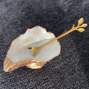 Peut inclure: Un plat en forme de coquille de couleur blanche et dorée avec une cuillère en forme de feuille dorée, rempli de cristaux de sel blanc.