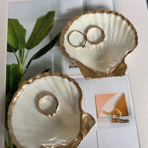 May include: Two white seashell-shaped ring dishes with gold trim. The dishes are on a white surface with a magazine in the background. One dish has two gold rings inside, and the other dish has one gold ring inside.
